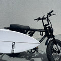 Black electric bike with a white surfboard attached against a gray wall